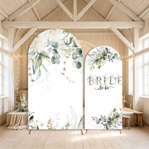 May include: Two arched backdrop panels featuring a white background with watercolor illustrations of green eucalyptus leaves, white flowers, and gold accents. The smaller panel displays the text "BRIDE to be" in a gold and black script font.