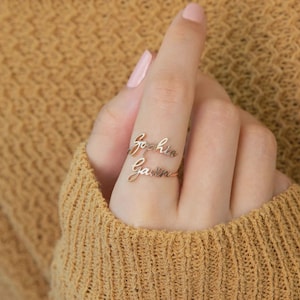 Può includere: Un anello in metallo color oro caratterizzato da due targhette sovrapposte con nomi in corsivo. La targhetta superiore riporta il nome Sophia e quella inferiore il nome Gavin. L'anello è realizzato con una struttura a fascia sottile e delicata.