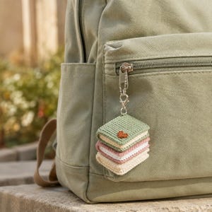 May include: A sage green canvas backpack featuring a silver metal zipper pull attached to a small, three-layered crochet book charm. The charm consists of stacked square books in sage green, dusty pink, and cream, with a small brown heart embroidered.