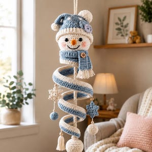 May include: A hanging crochet snowman decoration featuring a blue and white striped spiral body, a matching blue hat with a pom-pom, and a blue scarf with a small wooden button. The design includes dangling crochet stars and spheres.