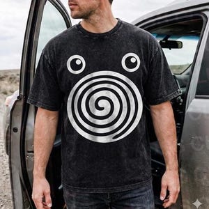 Könnte beinhalten: Ein anthrazitfarbenes T-Shirt in Acid-Wash-Optik mit einer großen weißen Spirale als Grafik auf der Brust, die unter zwei kreisförmigen weißen Augen platziert ist. Das Kleidungsstück hat einen Rundhalsausschnitt und kurze Ärmel mit Stoffstruktur.