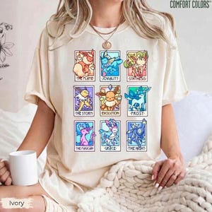 Könnte beinhalten: Ein cremefarbenes Kurzarm-T-Shirt mit einem Raster aus neun bunten Illustrationen fiktiver Kreaturen. Jedes Feld enthält eine Figur mit einer Beschriftung, darunter The Flame, Joviality, Synthesis, The Storm, Evolution, Frost, The Magician, Grace und The Night.