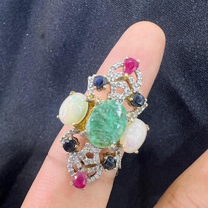 May include: A gold ring featuring a large central oval green emerald, flanked by two white opals. The setting is accented with small round dark blue sapphires, pear-shaped red rubies, and numerous tiny clear pavé-set stones in a decorative metal frame.