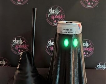 Puede incluir: Un enfriador de latas negro impreso en 3D con forma de fantasma y ojos ovalados de color verde brillante, coronado con una lata de aluminio plateada con el texto Contains 0% Juice y Nutrition Facts, junto a un soporte para pajitas de bruja.