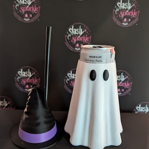 May include: A white ghost-shaped beverage can cooler with two black oval eyes, positioned next to a black witch hat-shaped accessory featuring a purple band and a tall black straw protruding from the centre of the hat's crown.