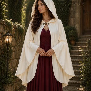 May include: A cream-colored hooded cloak featuring a metallic bronze-toned clasp at the neckline. The cloak is worn over a floor-length, sleeveless burgundy dress with a pleated bodice. The garment is made of a smooth, solid-colored fabric.