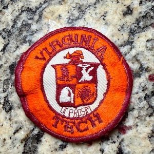 May include: A circular embroidered patch featuring a maroon and orange border. The centre displays a shield with four quadrants in maroon and white, topped with a small figure. The text VIRGINIA TECH and UT PROSIM are stitched in maroon.