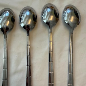 May include: Four polished stainless steel spoons arranged in a vertical row. Each spoon features a rounded bowl and a long, flat handle decorated with a series of horizontal and vertical etched lines that create a grid-like pattern along the stem.