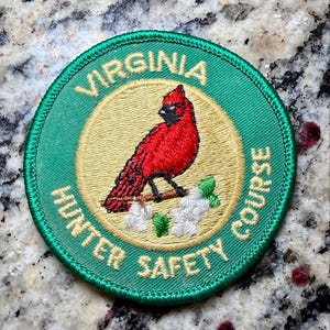 May include: A circular embroidered patch with a green border and a tan center. It features a red cardinal bird perched on a brown branch with white flowers and green leaves. The text VIRGINIA HUNTER SAFETY COURSE is stitched in tan lettering.