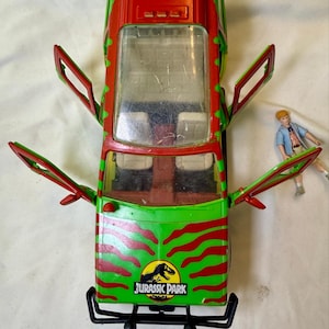 Könnte beinhalten: Ein Jurassic Park Spielzeugfahrzeug mit einer leuchtend grünen und roten Streifenlackierung, vier offenen Türen und einer klaren Windschutzscheibe. Auf der Motorhaube befindet sich ein kreisförmiges Jurassic Park Logo. Eine kleine blonde Figur steht daneben.