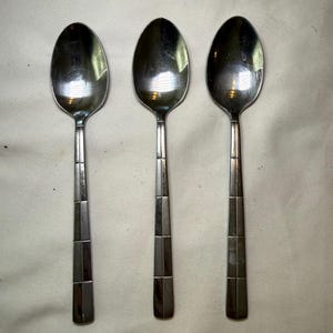 May include: Three identical silver-toned metal spoons arranged vertically. Each spoon features a smooth, oval-shaped bowl and a flat handle decorated with a series of horizontal and vertical etched lines that create a grid-like pattern along the stem.