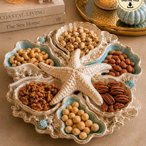 May include: A circular serving tray featuring five shell-shaped compartments arranged around a central starfish. The tray has a cream-colored exterior with light blue interior accents and small blue sea urchin details. It is filled with various nuts.