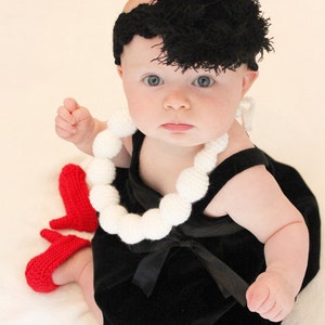 Könnte beinhalten: Ein Baby in einem schwarzen Samtkleid, einer weißen Perlenkette und einer schwarzen Perücke. Rote gestrickte Babyschuhe sind zu sehen. Das Bild enthält den Text "Sahrit eye for cuteness © 2016 All Rights Reserved."