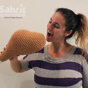May include: A woman is holding a large crocheted brown chicken leg. She is smiling and looking at the leg. The leg is made of brown yarn and has a white crocheted foot.