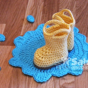 Puede incluir: Un par de botas de lluvia de crochet amarillo con un charco de crochet azul debajo. Las botas tienen una suela de crochet marrón.
