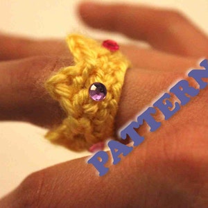 May include: A yellow crocheted crown ring with pink and purple rhinestones. The ring is worn on a finger.
