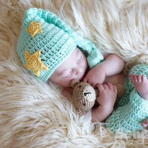 May include: A baby wearing a light blue crocheted hat with yellow stars and a matching light blue crocheted outfit is sleeping on a white fluffy blanket. The baby is holding a brown crocheted teddy bear.