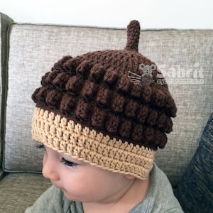 Op de afbeelding: Een bruine en beige gebreide eikel muts met een puntige top. De muts wordt gedragen door een baby.
