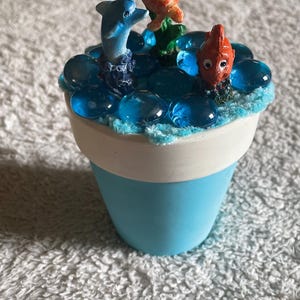 May include: A light blue flower pot with a white rim, filled with blue glass pebbles and small figurines of a blue dolphin, an orange sea turtle, and an orange fish. The top edge of the pot is decorated with a textured light blue material.