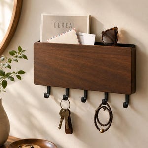 May include: A rectangular dark wood wall-mounted organizer featuring a top slot for mail and five black metal hooks underneath. The organizer holds a magazine labeled CEREAL, an envelope, and sunglasses, with keys and a bracelet hanging from the hooks.