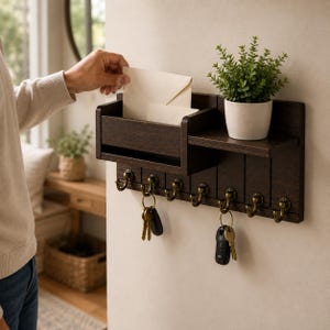 May include: A dark brown wooden wall-mounted organizer featuring a mail slot, a small shelf holding a potted green plant, and seven antique brass-toned metal hooks. Two sets of keys hang from the hooks, and a cream-colored envelope is placed inside.