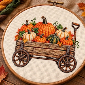 Peut inclure: Un projet de broderie au point de croix dans un tambour en bois rond représentant un chariot en bois rempli de citrouilles de différentes teintes orange, blanches et vertes, agrémenté de vignes vertes et de feuilles d'automne sur fond blanc.