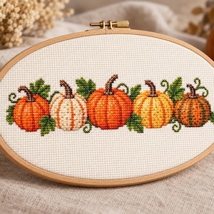May include: An oval wooden embroidery hoop containing a cross-stitch pattern on white fabric. The design features five pumpkins in varying shades of orange, yellow, and white, surrounded by green leaves and curling vines, centred within the frame.