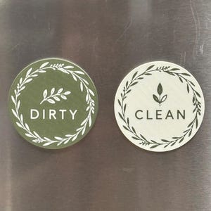 May include: Two circular magnets for a dishwasher. The left magnet is olive green with a white leaf wreath, a small leaf sprig, and the word DIRTY. The right magnet is off-white with a dark green leaf wreath, a small leaf sprig, and the word CLEAN.