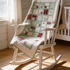 May include: A white wooden rocking chair draped with a patchwork quilt featuring a grid pattern of sage green, cream, and deep red fabric squares. The quilt has a repeating floral and geometric print, with a cream border featuring small dark dots.