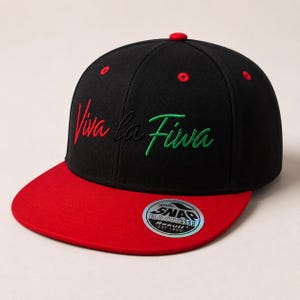 Könnte beinhalten: Eine schwarze Baseballkappe mit flachem rotem Schirm und einem roten Knopf oben. Die Vorderseite zeigt den gestickten Schriftzug Viva la Fiwa in roter, schwarzer und grüner Schreibschrift. Ein runder silberner Aufkleber klebt auf dem Schirm.