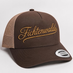 Könnte beinhalten: Eine braune Trucker-Cap mit Netzrückseite und festem Vorderteil. Das Wort Fichtenwalde ist in goldener Schreibschrift auf die Vorderseite gestickt. Ein runder Aufkleber mit dem Text The Authentic Yupoong Snapback befindet sich auf dem Schirm.