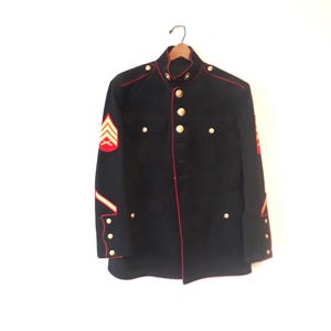 May include: A navy blue military jacket with red trim and gold buttons. The jacket has red and gold insignia on the sleeves and a red and gold chevron patch on the left sleeve. The jacket is hanging on a wooden hanger.