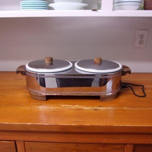 May include: A chrome and wood double electric chafing dish with two round white ceramic bowls. The chafing dish is on a wooden table.