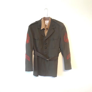 May include: An olive green military jacket with a matching belt and a tan collared shirt underneath. The jacket has red insignia on the sleeves. The jacket is hanging on a metal hanger against a white background.