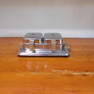 May include: A vintage chrome waffle iron with two separate compartments and wooden handles. The waffle iron is sitting on a wooden surface.