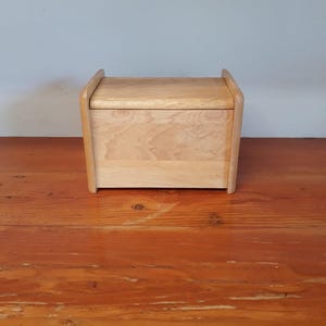May include: A light-colored wooden box with a lid. The box has a rectangular shape with rounded edges and a handle-like design on the top. The wood grain is visible, and the box sits on a wooden surface.