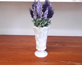 May include: A white, vintage-style vase with embossed floral designs holds a bundle of purple lavender. The vase has a pedestal base and a flared top. The arrangement sits on a wooden surface.