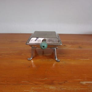 May include: A silver metal ashtray with a green plastic handle and four legs. The ashtray is on a wooden surface.