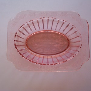 May include: A pink glass dish with a scalloped edge and a raised, oval design in the center. The dish has a delicate, floral pattern around the rim.