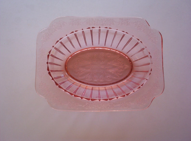 May include: A pink glass dish with a scalloped edge and a raised, oval design in the center. The dish has a delicate, floral pattern around the rim.