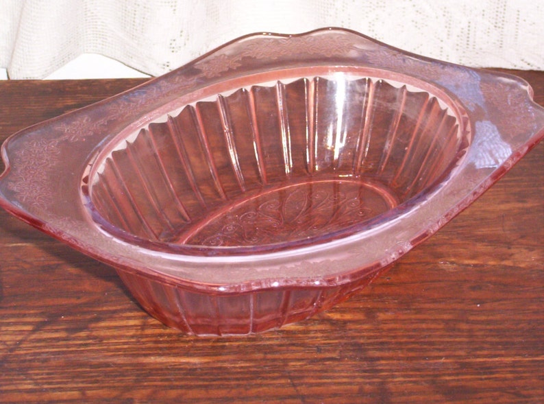 May include: A pink glass bowl with a fluted design and a scalloped edge. The bowl has a delicate floral pattern etched into the glass.
