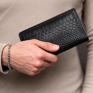 May include: A rectangular black wallet featuring a textured, embossed pattern that resembles reptile scales. The wallet has a slim, folded design with clean, stitched edges along the perimeter, showcasing a uniform and matte black finish.