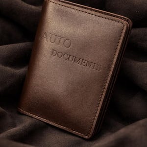 May include: A dark brown leather document holder featuring the embossed text AUTO DOCUMENTS on the front cover. The rectangular wallet has visible stitching along the edges and a smooth, matte finish, designed for organising vehicle paperwork.