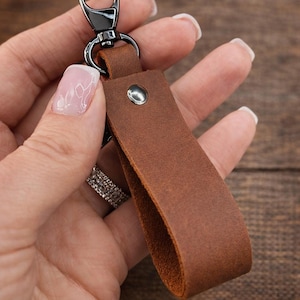 May include: A brown leather keychain strap featuring a smooth matte finish, secured with a single silver-toned metal rivet. The strap is attached to a black metal swivel lobster clasp, providing a functional and simple design for holding keys.