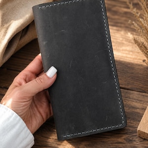 May include: A rectangular black leather wallet featuring a matte finish and visible light-colored stitching along the edges. The wallet has a slim, vertical orientation with rounded corners and a smooth, uniform surface texture throughout the front panel.