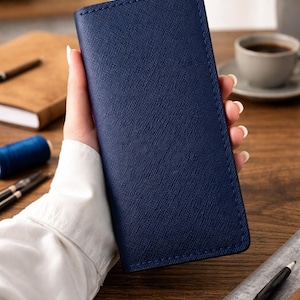 May include: A tall, rectangular navy blue wallet with a textured, cross-hatched surface finish. The wallet features clean, straight edges and matching navy blue stitching along the perimeter, presenting a slim and structured vertical design.