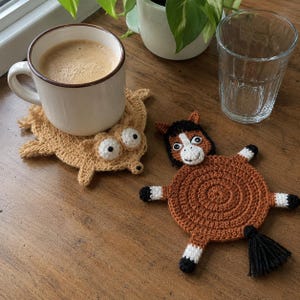 Pode incluir: Dois porta-copos de crochê feitos à mão em formato de animais. Um é uma criatura bege claro com olhos brancos grandes sob uma caneca de cerâmica branca com café. O outro é um cavalo marrom com crina preta, marcas brancas e cauda preta.