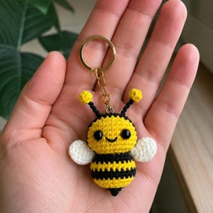 Pode incluir: Um pequeno porta-chaves de abelha feito em croché com um corpo às riscas amarelas e pretas, duas asas brancas e antenas pretas com pontas amarelas. A abelha tem dois olhos pretos redondos e um sorriso bordado, preso a um anel de metal dourado.