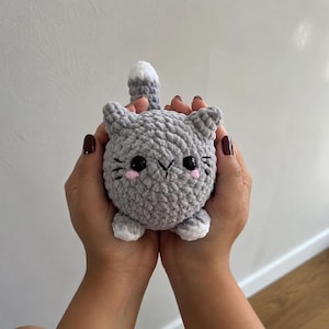 Pode incluir: Um peluche de gato redondo em croché cinzento com a ponta da cauda branca, patas brancas e orelhas pequenas. O gato apresenta olhos de plástico preto, um nariz preto bordado e bochechas cor-de-rosa, feito de fio macio e texturado.