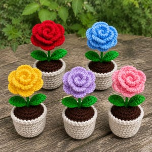 Pode incluir: Cinco flores de crochê em vasos nas cores vermelho, azul, amarelo, roxo e rosa. Cada flor apresenta pétalas em camadas, duas folhas verdes e terra marrom escura dentro de um pequeno vaso de crochê bege claro sobre uma superfície de madeira.